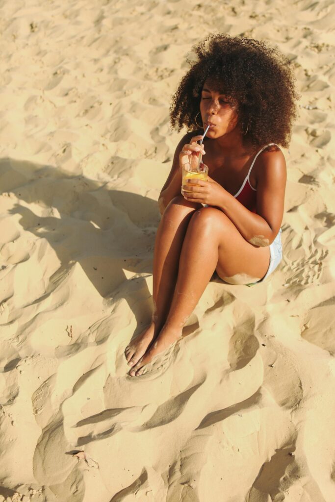 Relaxed woman with curly hair sipping a drink on a sunny sandy beach.